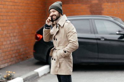 Close-up of a young, smiling, happy, laughing hipster with a beard in a raincoat and hat,