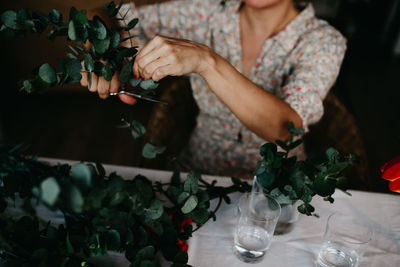 Unrecognizable female with scissors cutting stems of plant with lush green foliage while sitting at table with glasses at home
