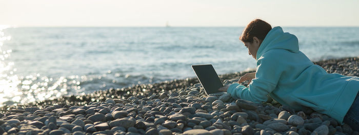 Caucasian woman working on a laptop on a pebbly seashore.