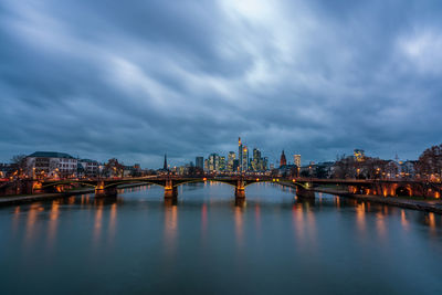 Illuminated bridge over river by buildings against sky at dusk