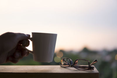 Cropped hand holding mug by eyeglasses on table 