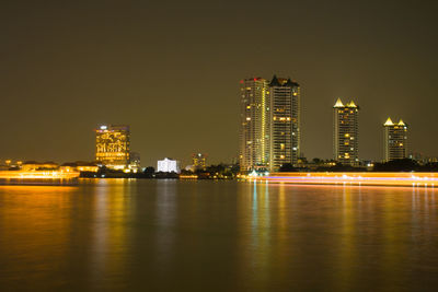 Illuminated buildings by river against sky at night