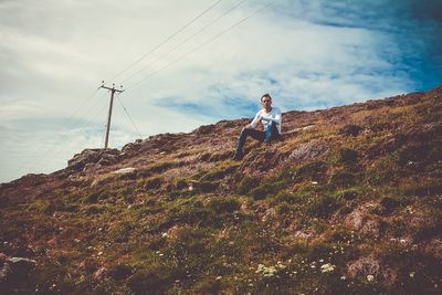 Low angle view of man sitting on mountain against cloudy sky