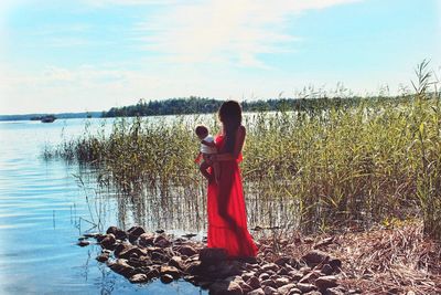 Full length of mother with baby standing by lake against sky during sunny day