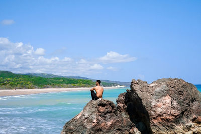 Man sitting on rock by sea against sky