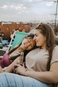 Smiling young woman holding beer bottle sitting by female friends with eyes closed on rooftop