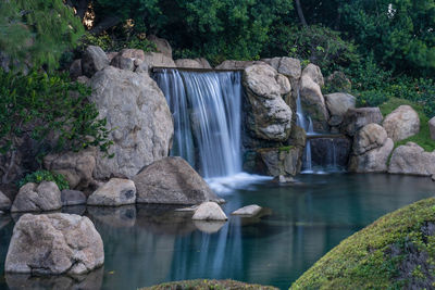 Peacefull waterfall is gently flowing in the park on a sunny day with blue waters and green foliage