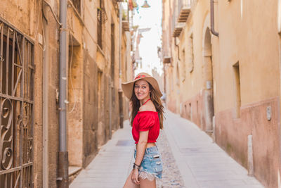 Smiling young woman wearing hat while standing in alley amidst buildings