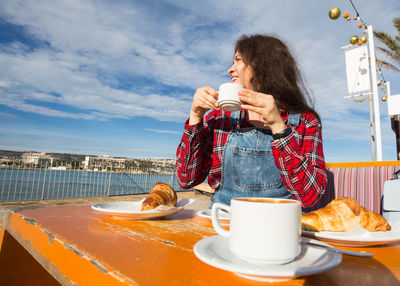 Young woman holding drink sitting on table against the sky