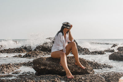 Full length of woman sitting on rock at beach against clear sky