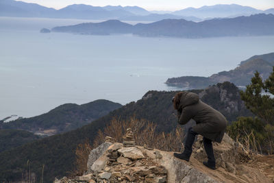 Side view of mature woman photographing sea while crouching on mountain against sky