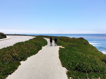 Rear view of people walking on road next to sea against clear sky