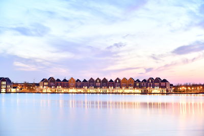 Lake by illuminated buildings against sky during sunset