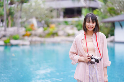 Portrait of woman holding camera standing by swimming pool