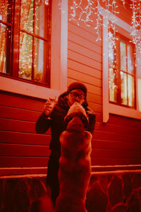 Man holding wineglass while standing by illuminated house with dog