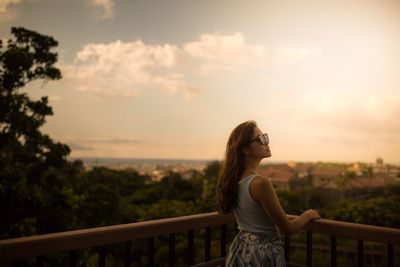 Woman standing by railing against sky during sunset