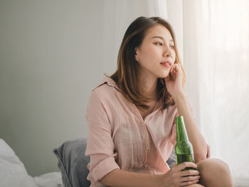 Thoughtful young woman looking away while holding bottle at home