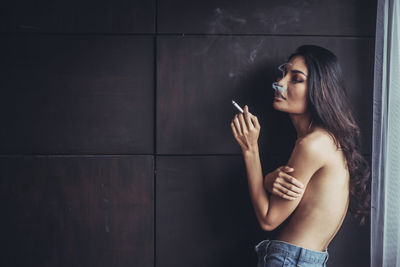 Shirtless young woman smoking cigarette against wall at home