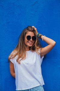 Portrait of smiling young woman in sunglasses standing against blue wall