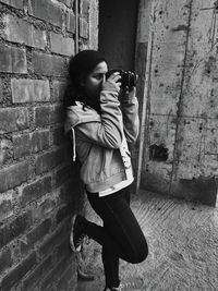 Full length of young woman photographing through camera while standing against wall