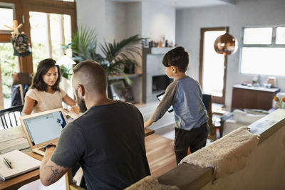 Rear view of man working on laptop while family at dining table in house