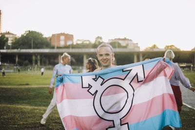 Portrait of happy non-binary person standing with transgender symbol on flag at park