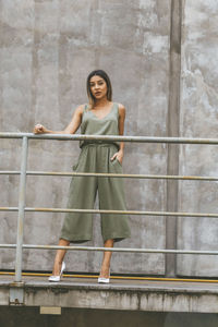 Full length of fashionable young woman standing by railing against concrete wall