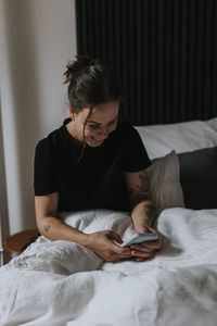 Smiling young woman sitting in bed and using cell phone