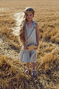 Girl child in a dress and a wreath on her head stands on a mown field of wheat at sunset in summer