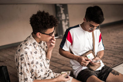 Man smoking cigarette by male counting paper currency while sitting on land