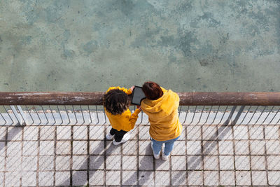 Top view of unrecognizable females in yellow clothes standing near railing in city and browsing tablet together