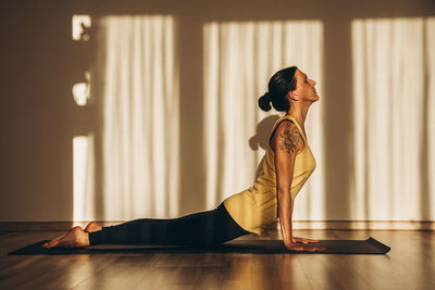  woman practicing yoga asana on black sport mat in a room with sunset or sunrise light with shadows.