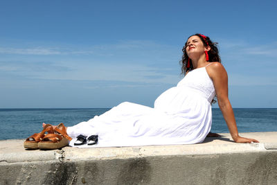 Young pregnant woman lying on seawall against sky with a pair of baby shoes  and her own next to her 