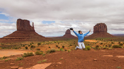 Portrait of cheerful woman with arms outstretched jumping on land against cloudy sky