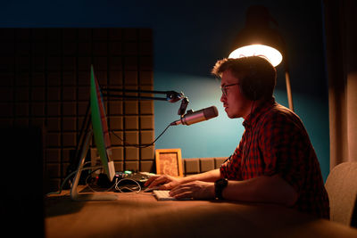 Side view of focused young male in checkered shirt and eyeglasses using computer and speaking in mic while recording podcast in dark studio