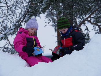 Cute kids reading book while sitting on snow during winter