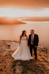 Rear view of bride and bridegroom standing at beach against sky during sunset