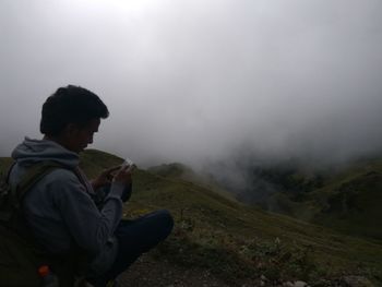 Man using mobile phone while sitting at mountain during foggy weather