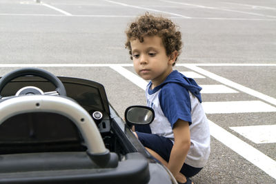 Curly-haired boy, looking attentively at the parked electric kiddie car.