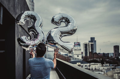 Rear view of man holding number 32 balloons on terrace in city