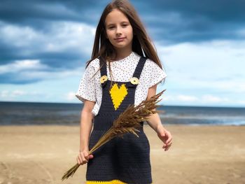 Portrait of girl holding plants standing at beach against sky