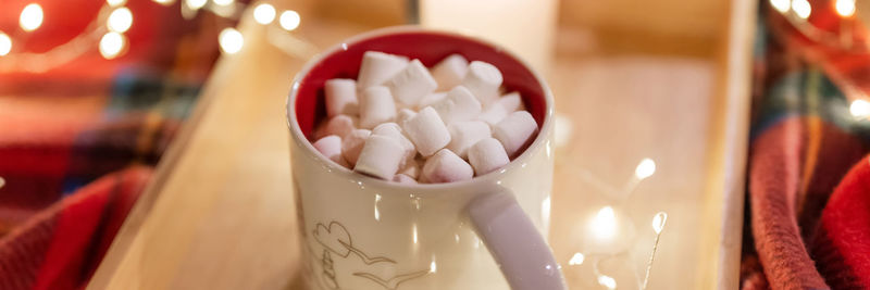 Cup of hot chocolate and marshmallow and burning candle on tray and a garland glowing lights