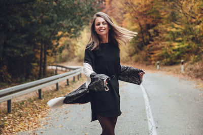 Portrait of smiling woman standing on road against trees during autumn