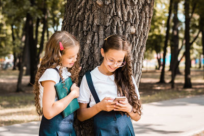Cheerful pupils in school uniform with a book looking at a smartphone in the park on a warm day
