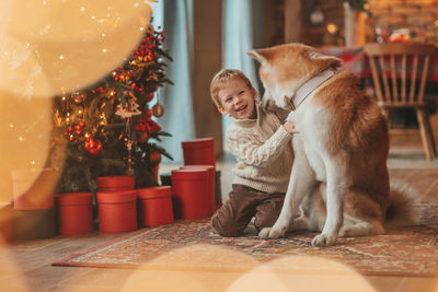 Candid authentic happy little boy in knitted beige sweater hugs dog with bow tie at home on xmas