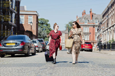 Two friends walking on the street with wheeled luggage and shopping bag looking at each other, liverpool, uk