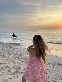 Rear view of woman standing at beach against sky during sunset