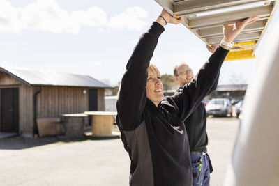 Smiling female electrician getting ladder with help of male coworker from van on sunny day