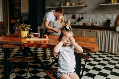 A little girl is holding a mug drinking tea and her parents are kissing from behind in the kitchen 