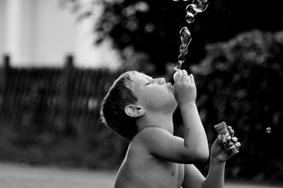 Shirtless boy blowing bubbles against tree